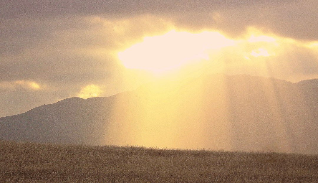Sunlight streams through gray clouds over rolling green hills after rain.